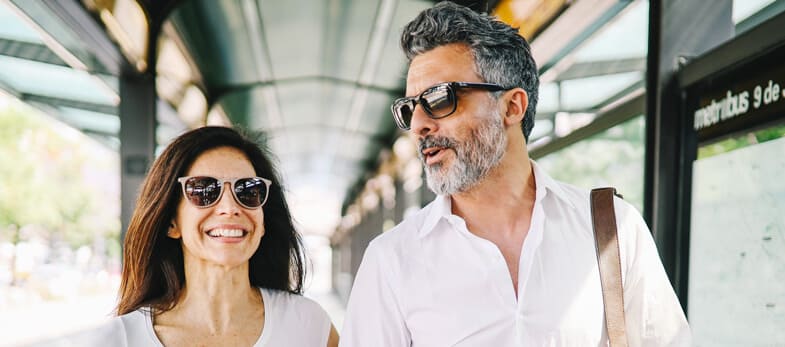 Dental Implants Wimbledon - A joyful middle-aged couple walks together, smiling confidently while enjoying a sunny day. Both are wearing sunglasses and casual white clothing, exuding a relaxed and happy demeanor. This image is associated with Ridgway Dental, a dental clinic in Wimbledon, highlighting the positive impact of dental implants provided by the clinic. The couple's bright smiles suggest successful dental treatments, emphasizing the clinic's expertise in restoring dental health and aesthetics. The background features an urban setting with a covered walkway, indicating a lively and vibrant area. This scene underscores Ridgway Dental's commitment to enhancing patients' lives through high-quality dental care in Wimbledon.