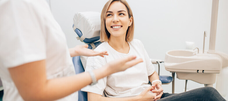 Specialist dentists Wimbledon - A young woman, seated comfortably in a dental chair at Ridgway Dental, a dental clinic in Wimbledon, smiles engagingly during a consultation with a specialist dentist. The dentist, partially visible, gestures while discussing treatment options, illustrating a patient-centered approach. The clinic's modern and well-equipped setting, with clean, bright interiors and essential dental tools in view, reinforces Ridgway Dental's commitment to providing top-notch dental care. This image highlights the friendly, professional environment that Ridgway Dental is known for, ensuring a pleasant and reassuring experience for every patient.