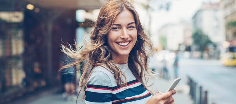 tooth coloured braces wimbledon - A young woman is smiling brightly while holding a smartphone, standing on a bustling street. Her wavy hair flows in the breeze, and she is dressed in a stylish, casual outfit. The background features an urban setting with blurred pedestrians and buildings, indicating a lively atmosphere. This image represents the vibrant and friendly community around Ridgway Dental, a dental clinic in Wimbledon, where patients can receive treatments such as tooth-coloured braces. The woman’s confident and radiant smile showcases the positive results of dental care available at Ridgway Dental, emphasizing the clinic's expertise in enhancing smiles with discreet orthodontic solutions.
